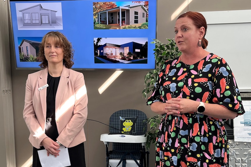 Two women stand in front of a screen displaying houses, giving a presentation.