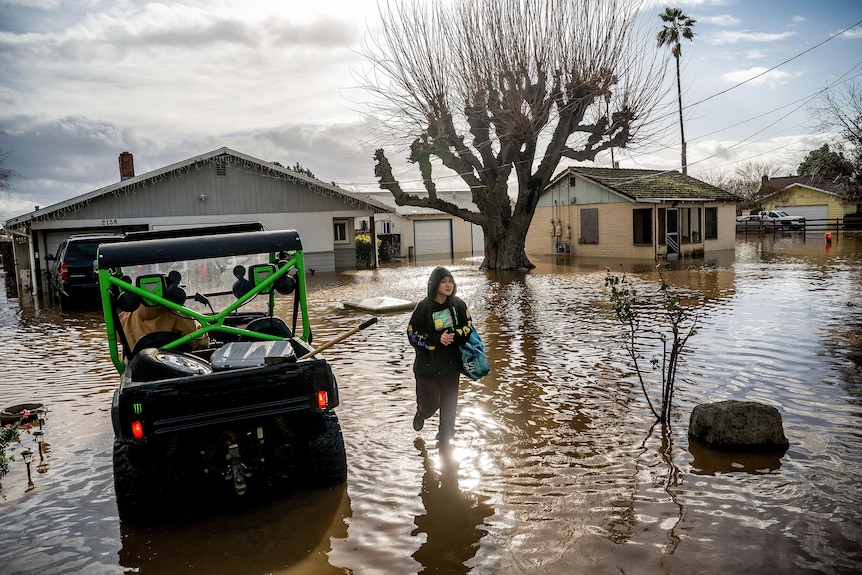 A person walking through flooded water next to a small ute in front of houses
