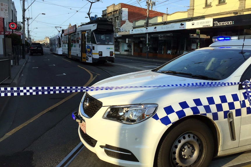 Two trams sit on the road behind police tape and a police car.