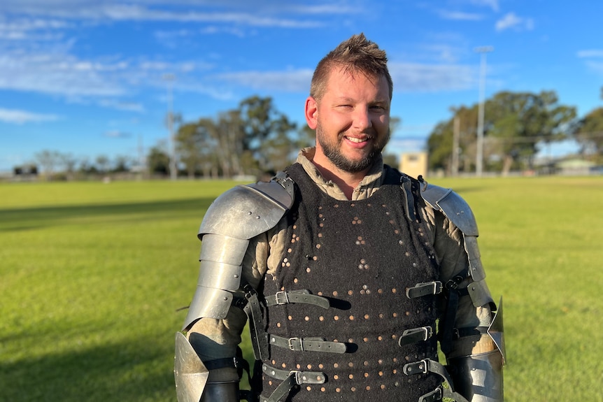 Smiling man wears silver armour on shoulders, arms, a black material chest piece, an olive green undershirt, in a large field.