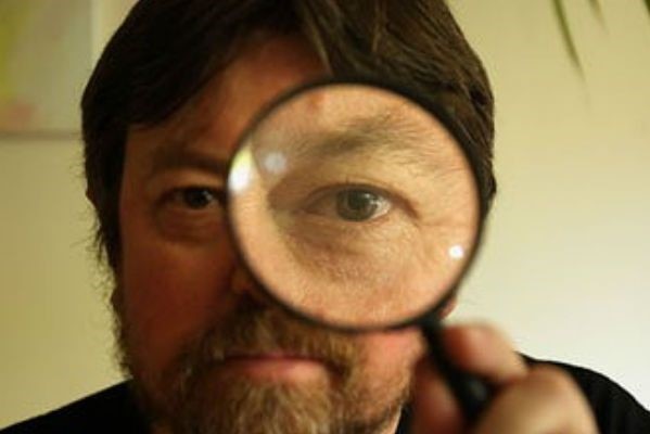 A man with a beard holding a magnifying glass which enlarges his left eye.