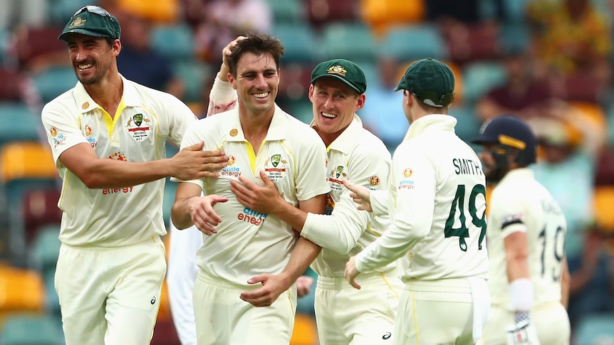 A group of smiling Australian men's cricketers hug and pat the bowler after a wicket during a Test match.
