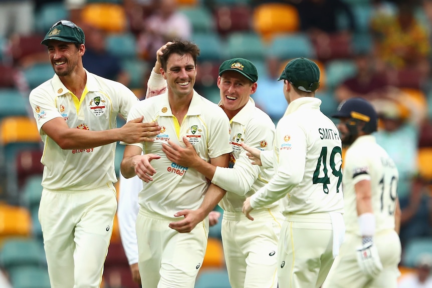 A group of smiling Australian men's cricketers hug and pat the bowler after a wicket during a Test match.