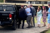 Michael McCormack helped by officials into a black car as people in floral headdresses watch on. 