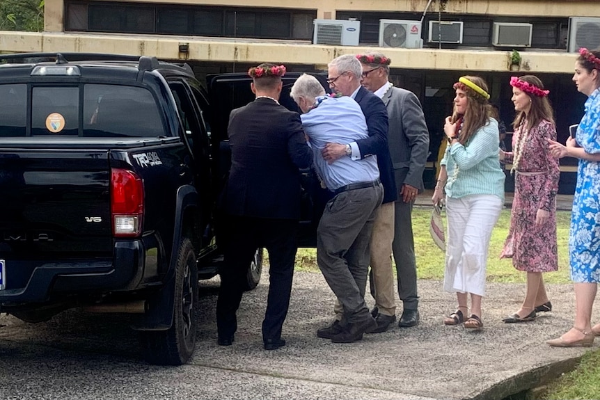 A man in business attire, unsteady on his feet, is helped into a vehicle by several people.