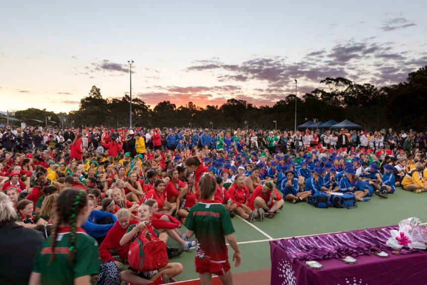 Thousands of netball players, coaches and officials dressed in team uniforms stand on netball courts at dusk.