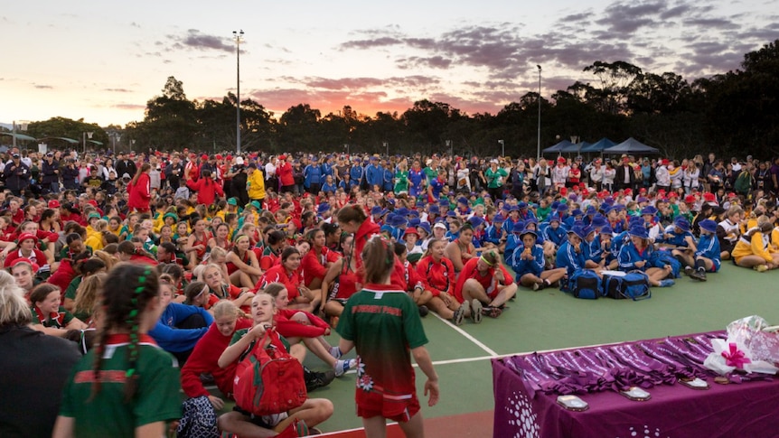 Thousands of netball players, coaches and officials dressed in team uniforms stand on netball courts at dusk.