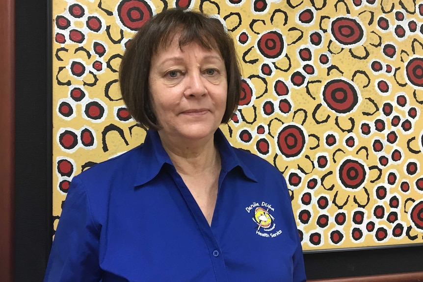A woman in a blue collared shirt stands in front of an Indigenous artwork.