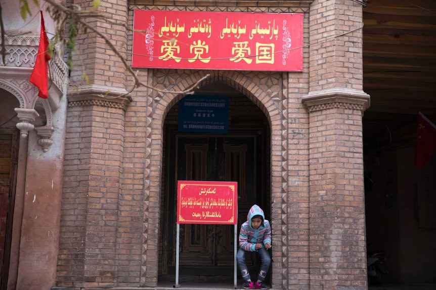 A child rests near the entrance to a mosque where a banner in red reads 