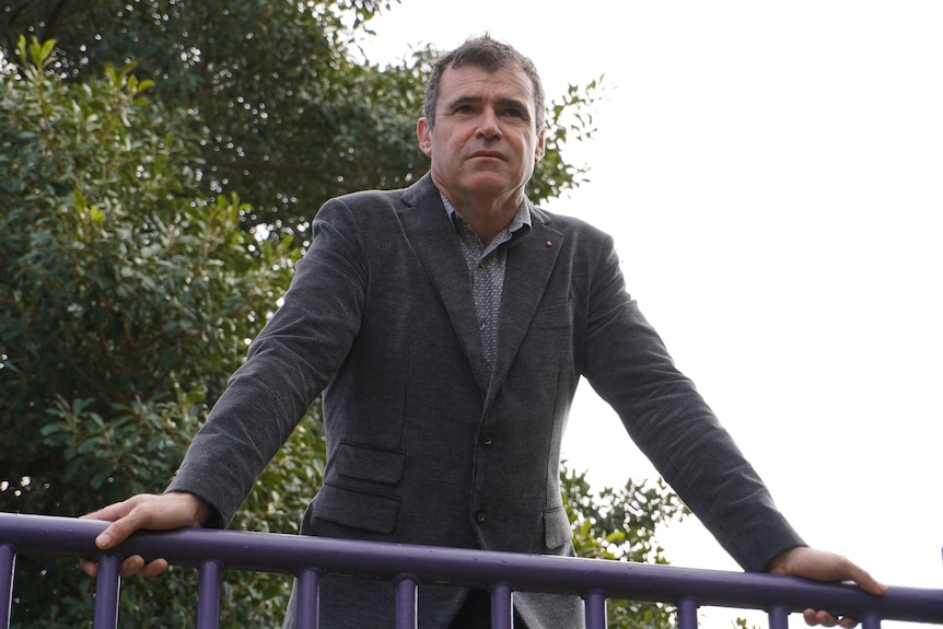 A man leans up against a railing outside looking serious as he gazes into the distance.