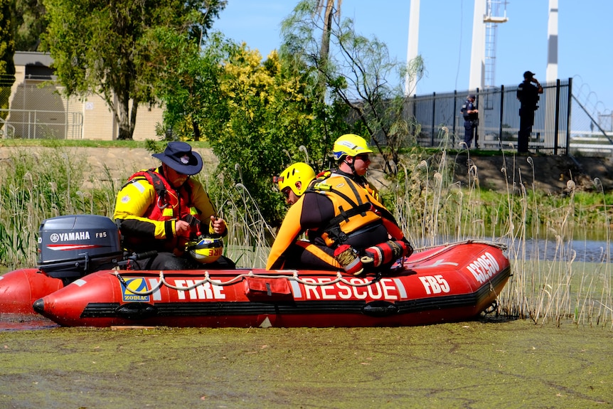 Three men in a red emergency rescue boat float along a green river while police on the banks look at the river 