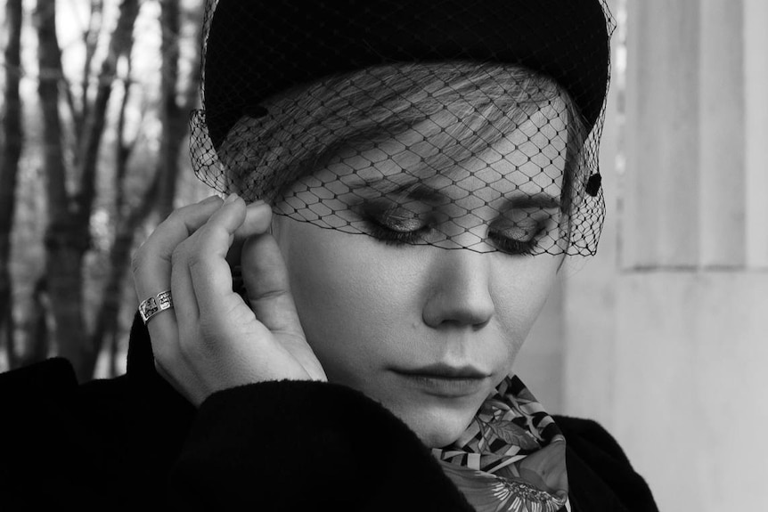 A black and white image of a young woman posing in a hat with black netting over her eyes