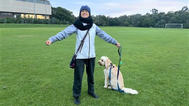 Small woman standing on green soccer pitch dressed in winter clothes holding arms out with dog on lead.