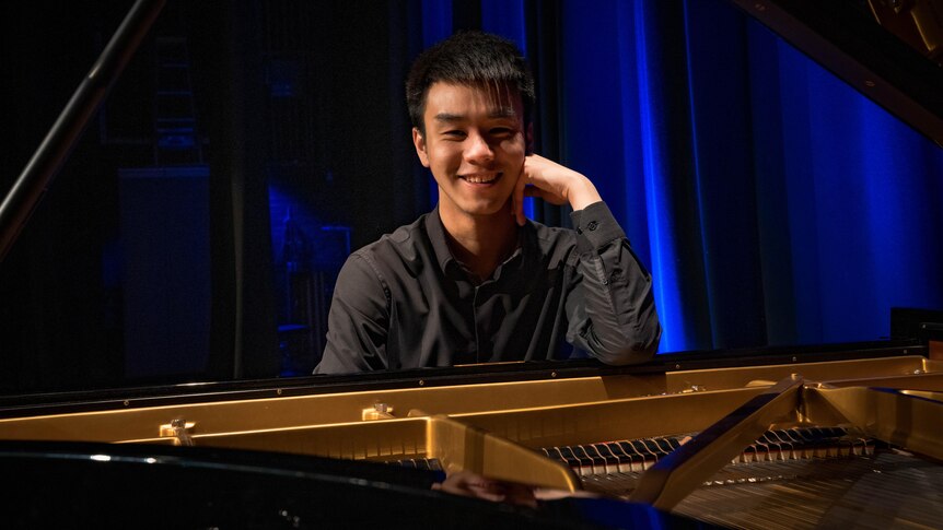 Pianist Alex Zhang sits behind a grand piano with the lid up. One elbow rests on the music stand and he is smiling.