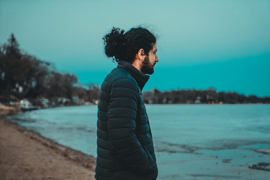 A man wears a puffer jacket with curly long hair tied back while standing with back to camera looking out to ocean 