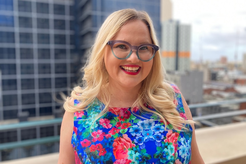 Sarah Moran, dressed in a vibrant, colourful top and wearing glasses, stands on a CBD rooftop.