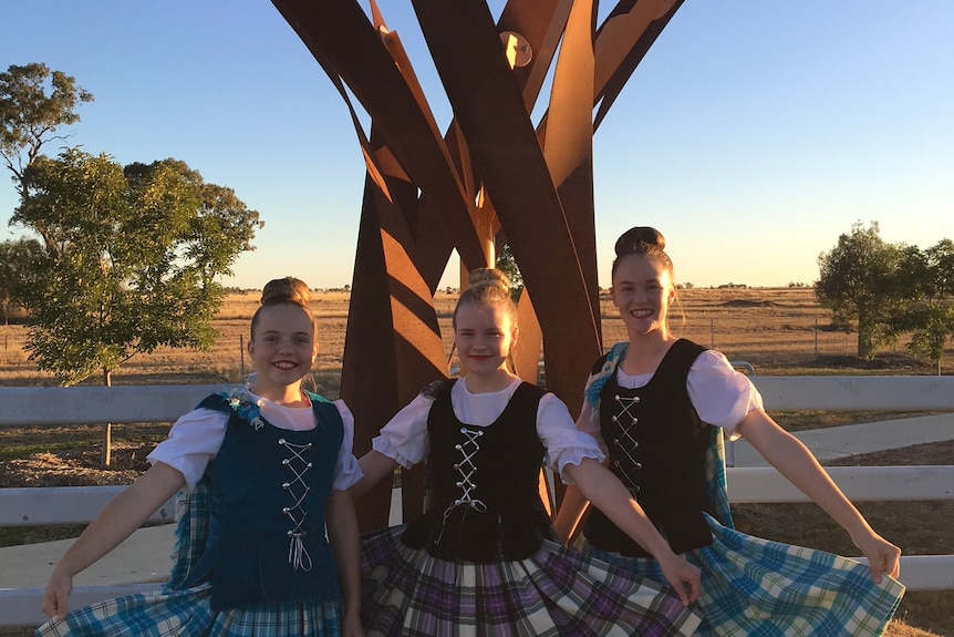 Three highland dancers in costume holding out their skirts and smiling in front of a rust coloured sculpture.