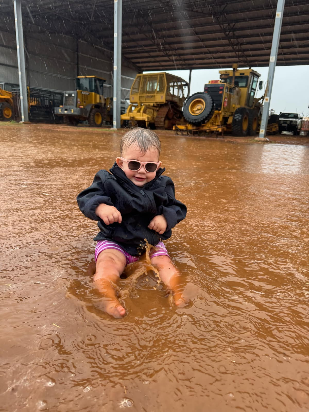 Child sits in rainwater smiling at camera at Mount Doreen homestead with shed in background with farm machinery