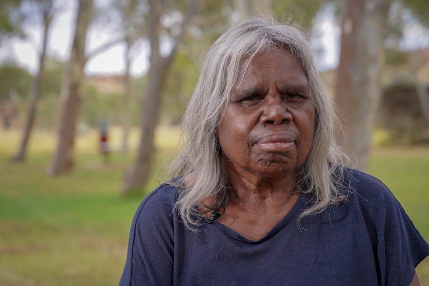 Older Indigenous woman wearing a navy shirt outside with trees behind her.