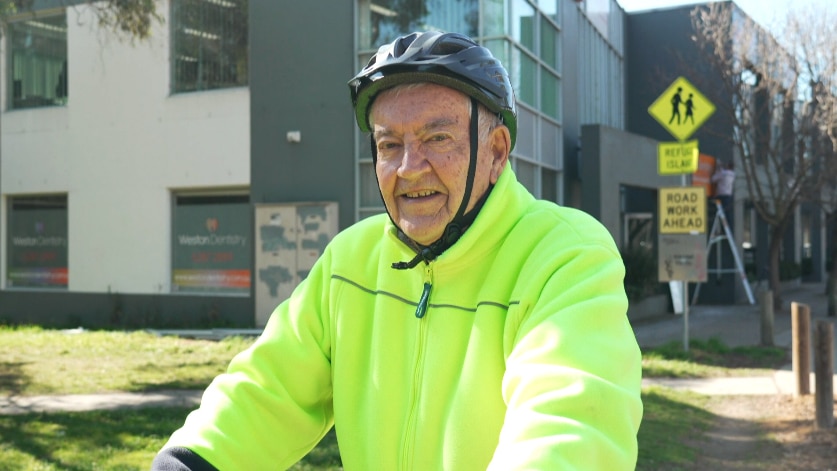 Ken wears high vis and a bike helmet, standing behind his bike on a street.