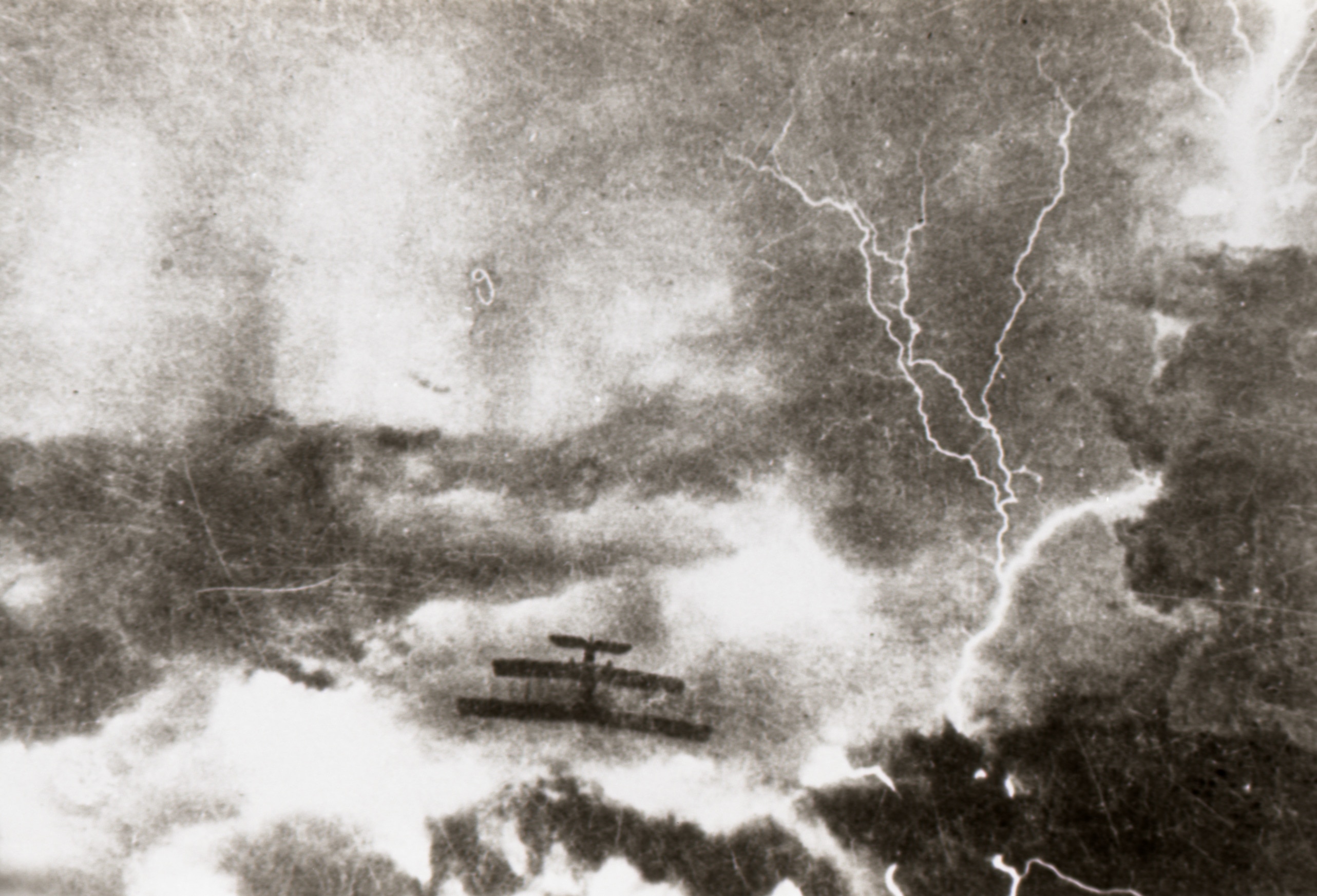 An airplane flies through clouds and lightning.  The photo is old and in black and white. 