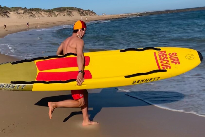 A young man runs into ocean carrying surf life saving rescue paddle board