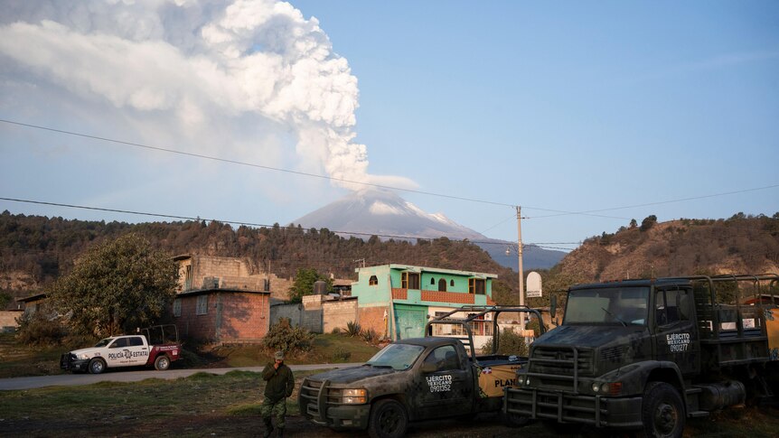 A white column of smoke rises from a volcano seen in the distance behind some Mexican buildings.