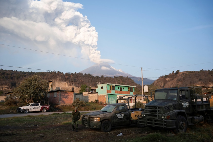 A white column of smoke rises from a volcano seen in the distance behind some Mexican buildings.