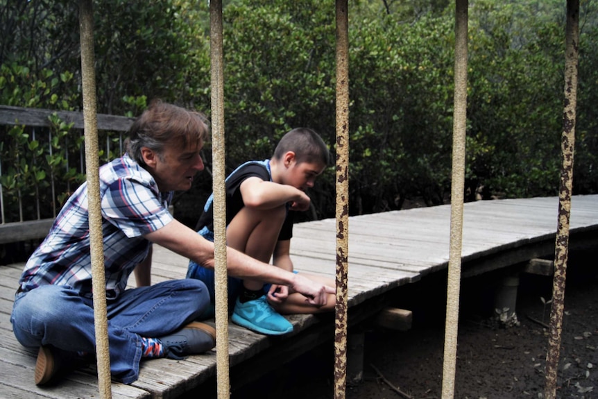 A man and a boy sit on a bridge, pointing to the creek below.