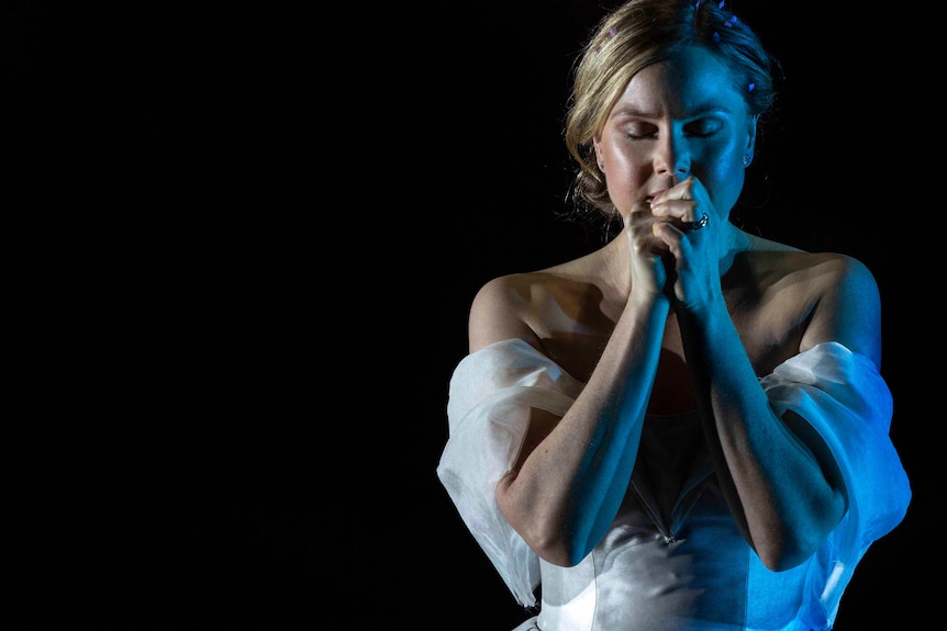 White woman with blonde hair elegantly pulled back wears white wedding dress and clasps hands together in a prayer.