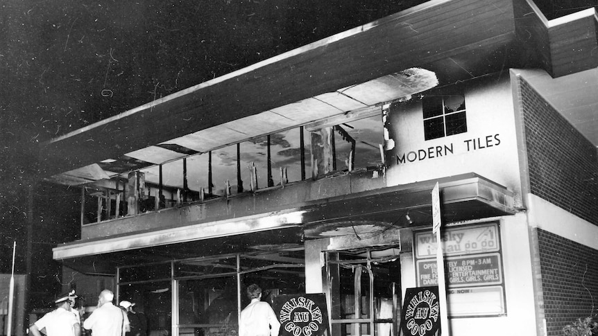 Black and white picture of police and onlookers standing outside a burnt out Whiskey Au Go Go nightclub