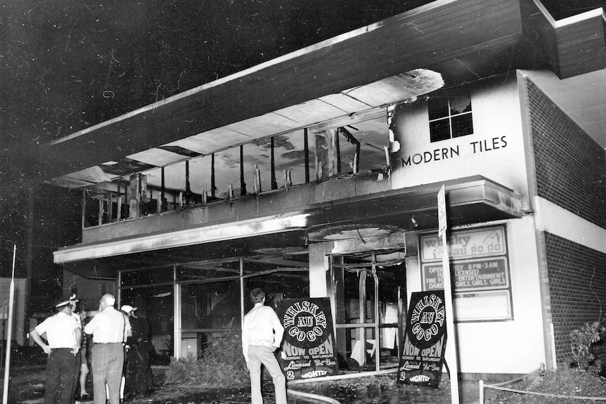 Black and white picture of police and onlookers standing outside the burnt-out Whiskey Au Go Go nightclub