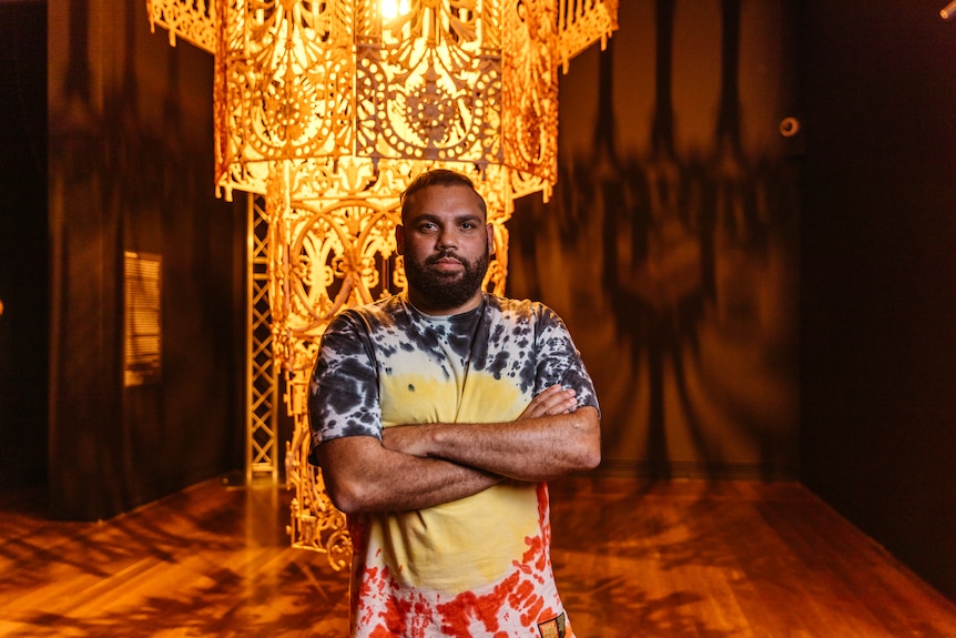 Warrang man with short dark hair and beard wears tie-dyed Aboriginal flag print shirt and stands before large yellow chandelier
