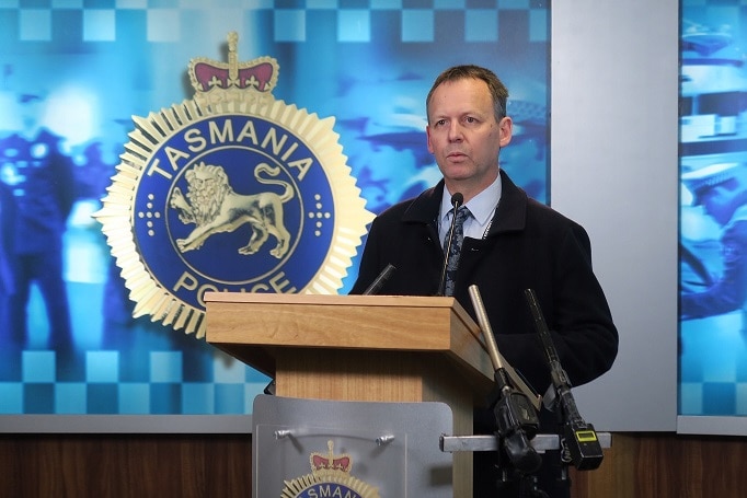 A man in a suit stands at a podium in front of a Tasmania Police logo