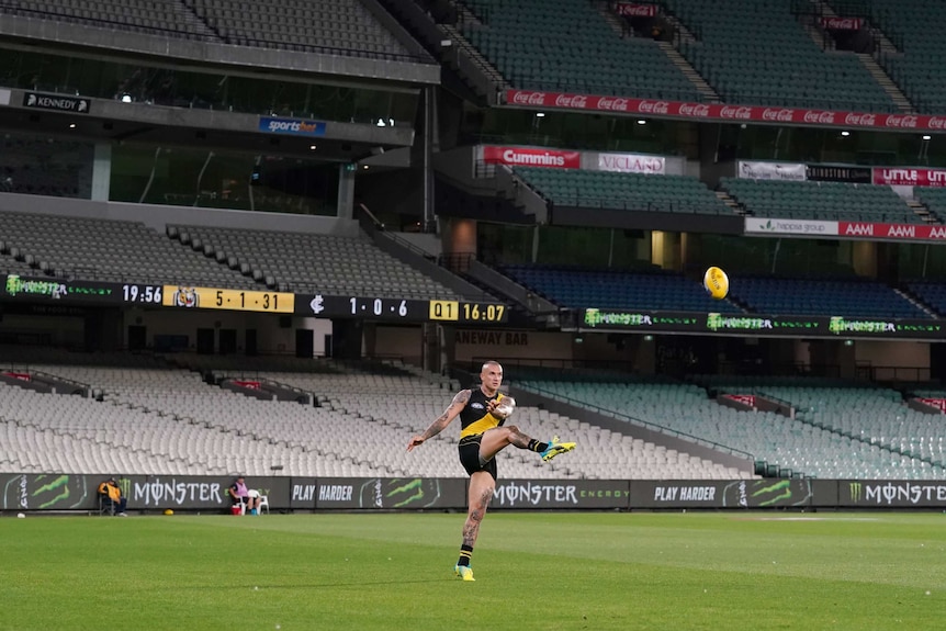 A male Richmond AFL player kicks the ball in front of an empty grandstand at the MCG.