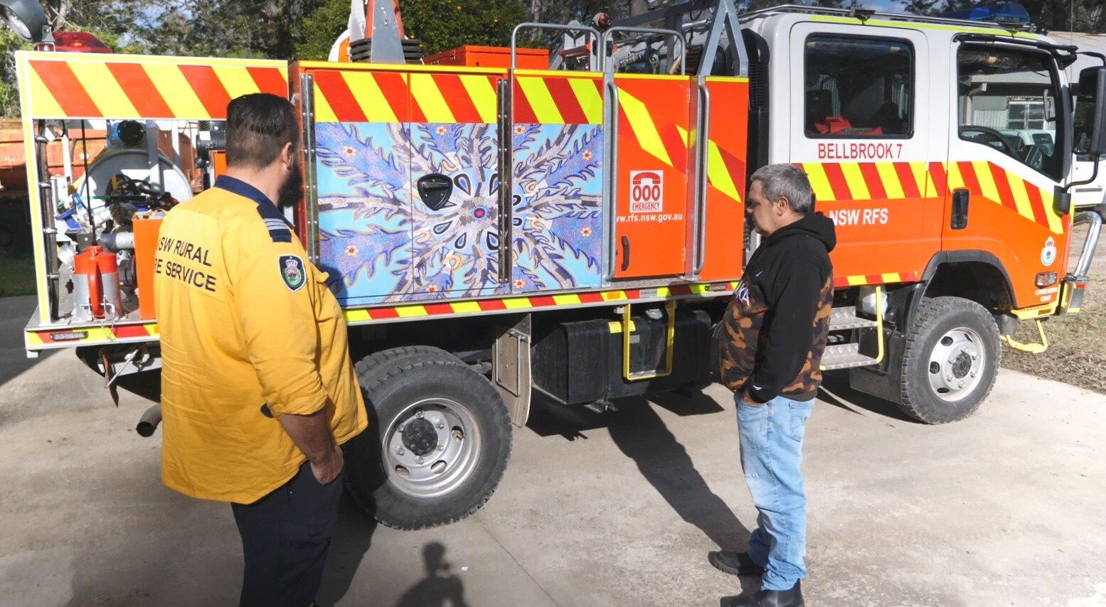 An Aboriginal man stands with a firefighter looking at an Indigenous artwork in blue tones, on the side of a red fire truck.