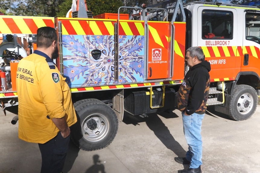 An Aboriginal man stands with a firefighter looking at an Indigenous artwork in blue tones, on the side of a red fire truck.