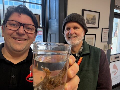 Two men smiling. One holds glass coffee cup with spotted handfish in it towards camera.