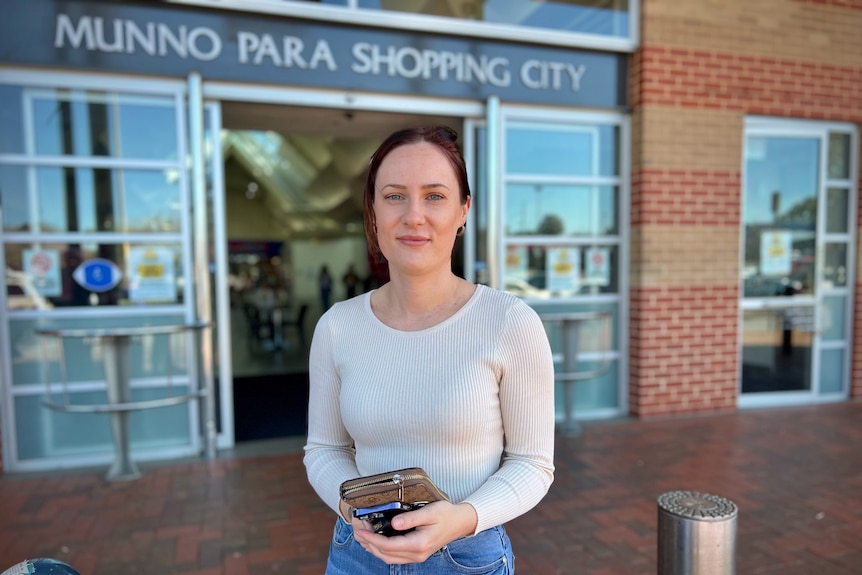 Una mujer con el pelo recogido y un suéter blanco parada frente a un centro comercial sonriendo a la cámara.