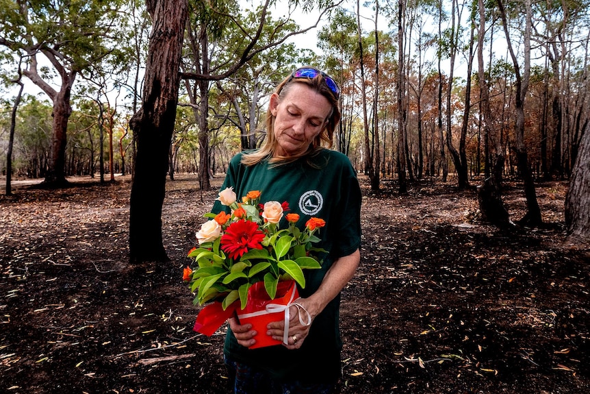 A woman looks down holding a box of flowers, burned trees behind her