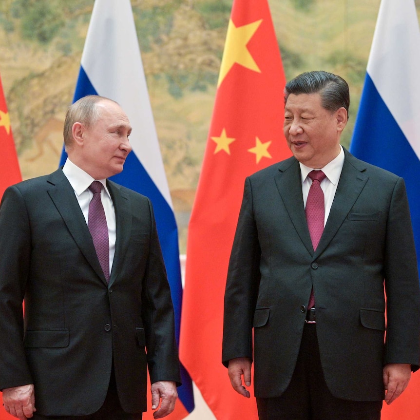 Two men in suits, Vladimir Putin and Xi Jinping, standing in front of flags.