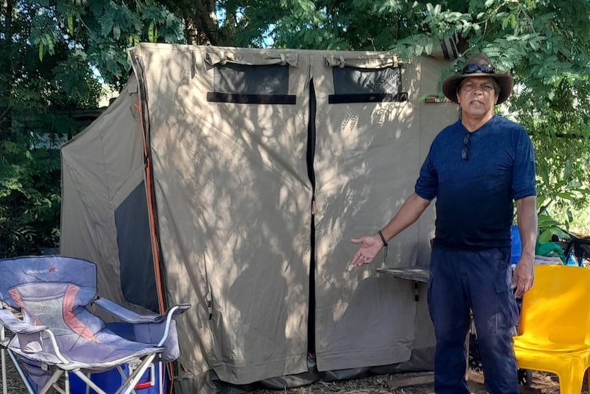 Peter is wearing a blue shirt and blue pants standing in front of the brown tent he now sleeps in