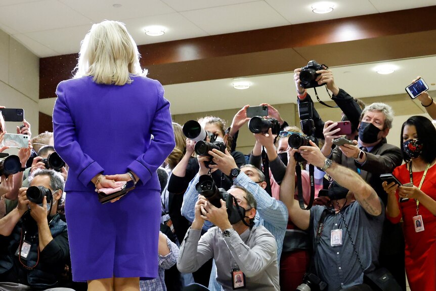 A  blonde woman in a purple skirt and blazer faces a mob of reporters with cameras 