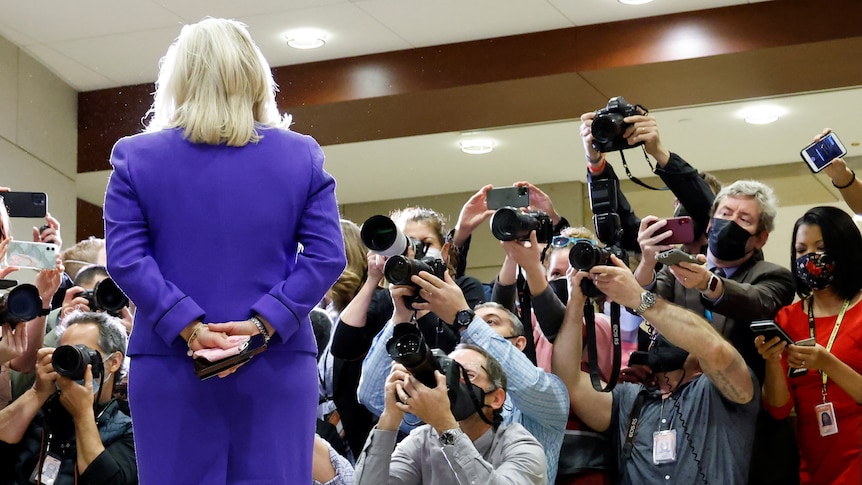 A  blonde woman in a purple skirt and blazer faces a mob of reporters with cameras 