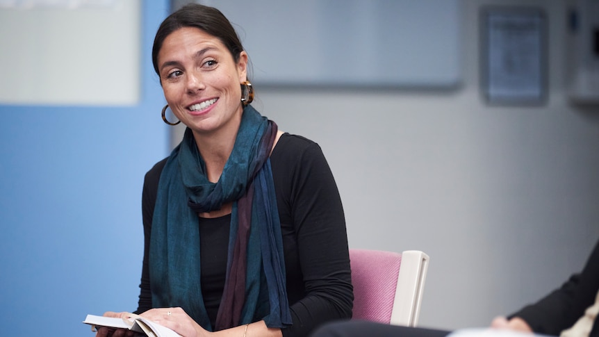 A woman wearing a scarf with a book open on her lap smiles. 