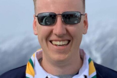 A smiling young man in sunglasses and a woggle standing in an alpine area.