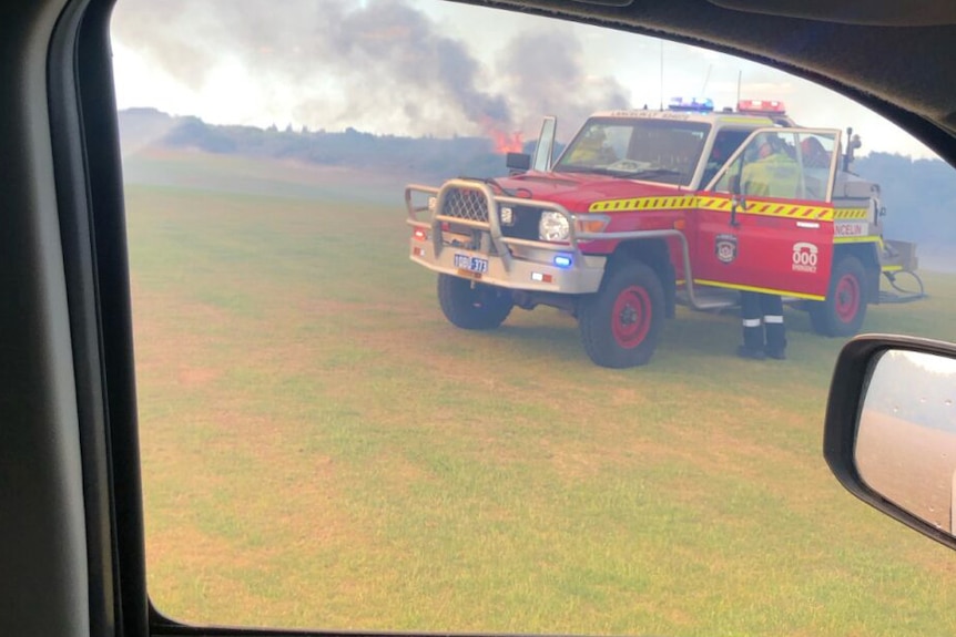 A fire truck on a golf course pictured through the window of a car, with a bushfire visible behind it.