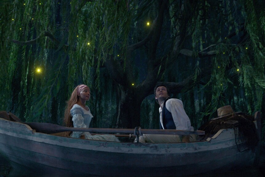 Halle Bailey as Ariel and Jonah Hauer-King as Prince Eric in The Little Mermaid. They're sitting in a boat, looking up at lights