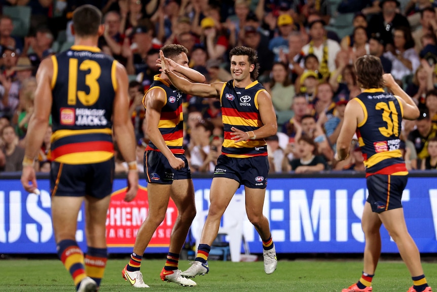 Josh Rachele points skyward and smiles as Crows teammates run to join him