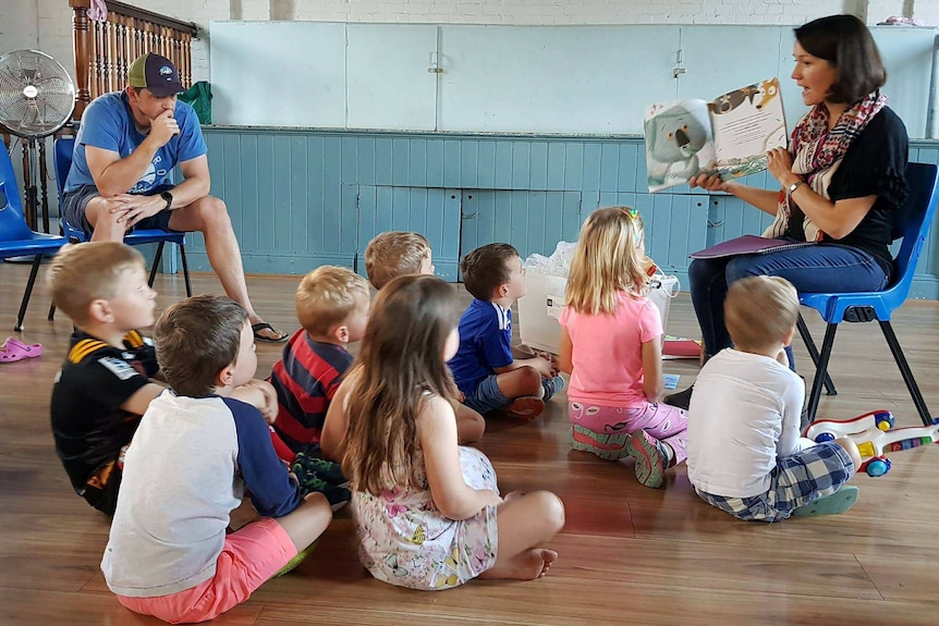 A woman reads a story book to a group of children sitting on the floor